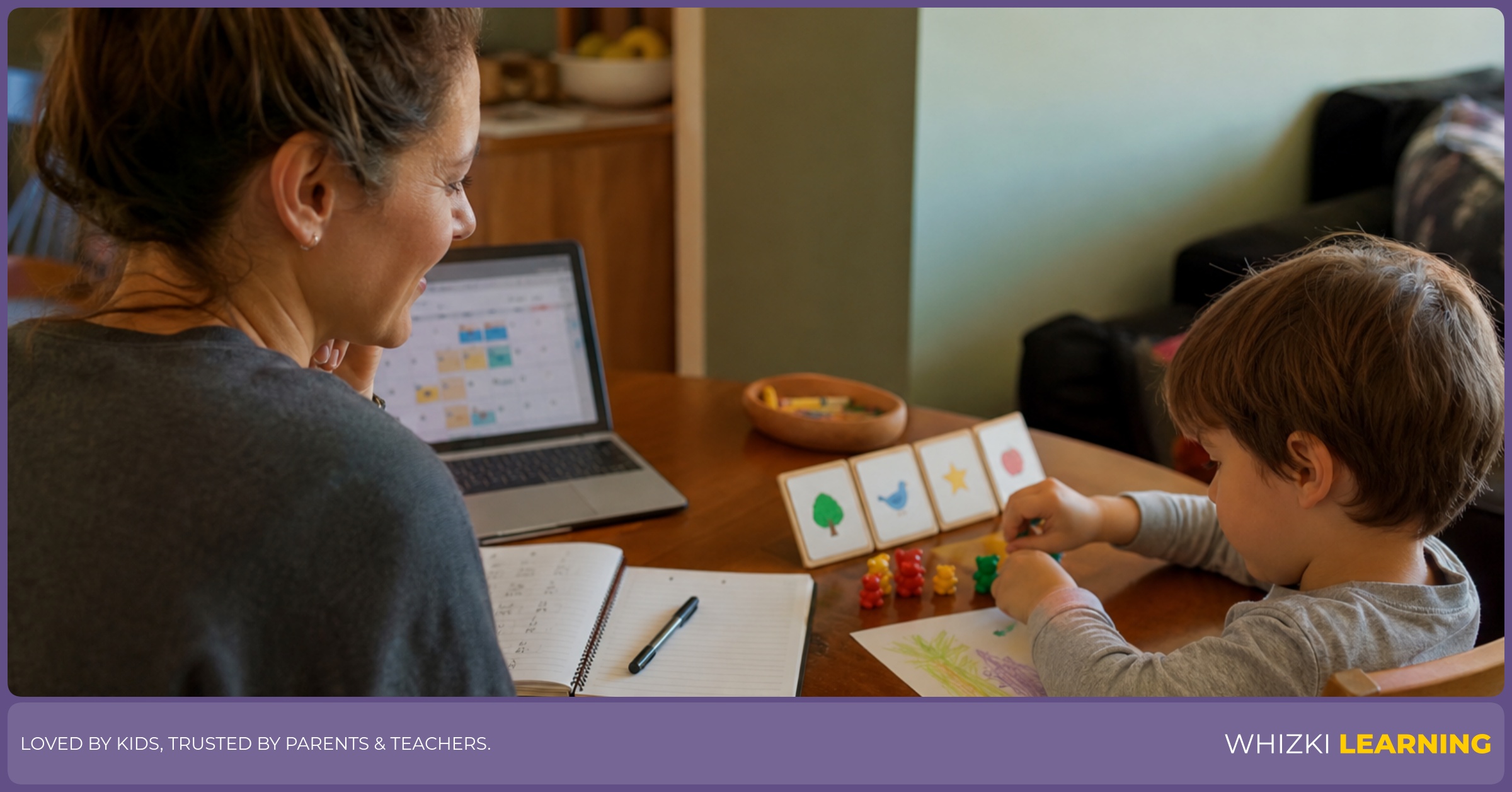 A parent glances at an adult online planner while a young child works with crayons and paper at the kitchen table.