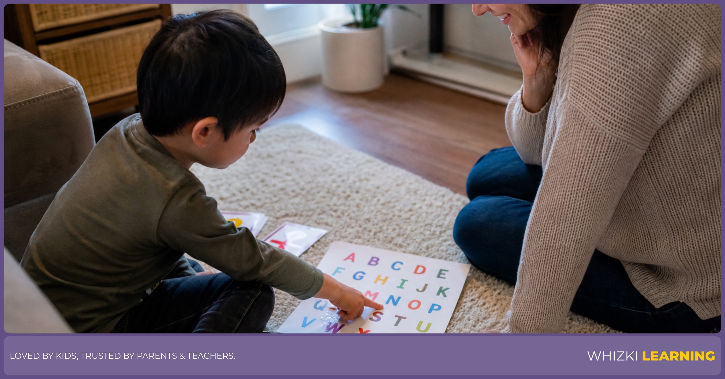 A caregiver watches a child trace a printed alphabet page with one finger on the living-room floor.