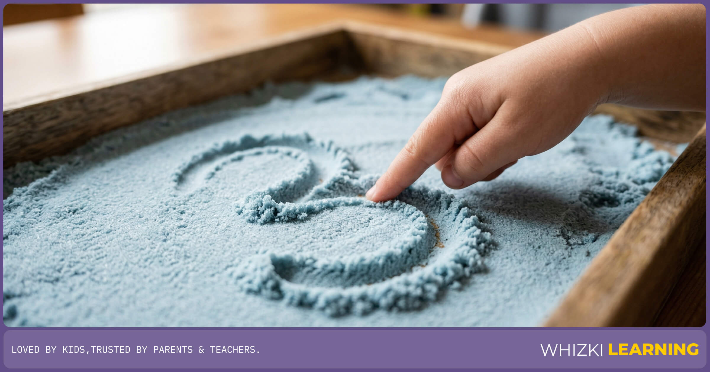 A close-up of a preschooler's hand drawing a number three in a shallow tray of kinetic sand to correct reversals.