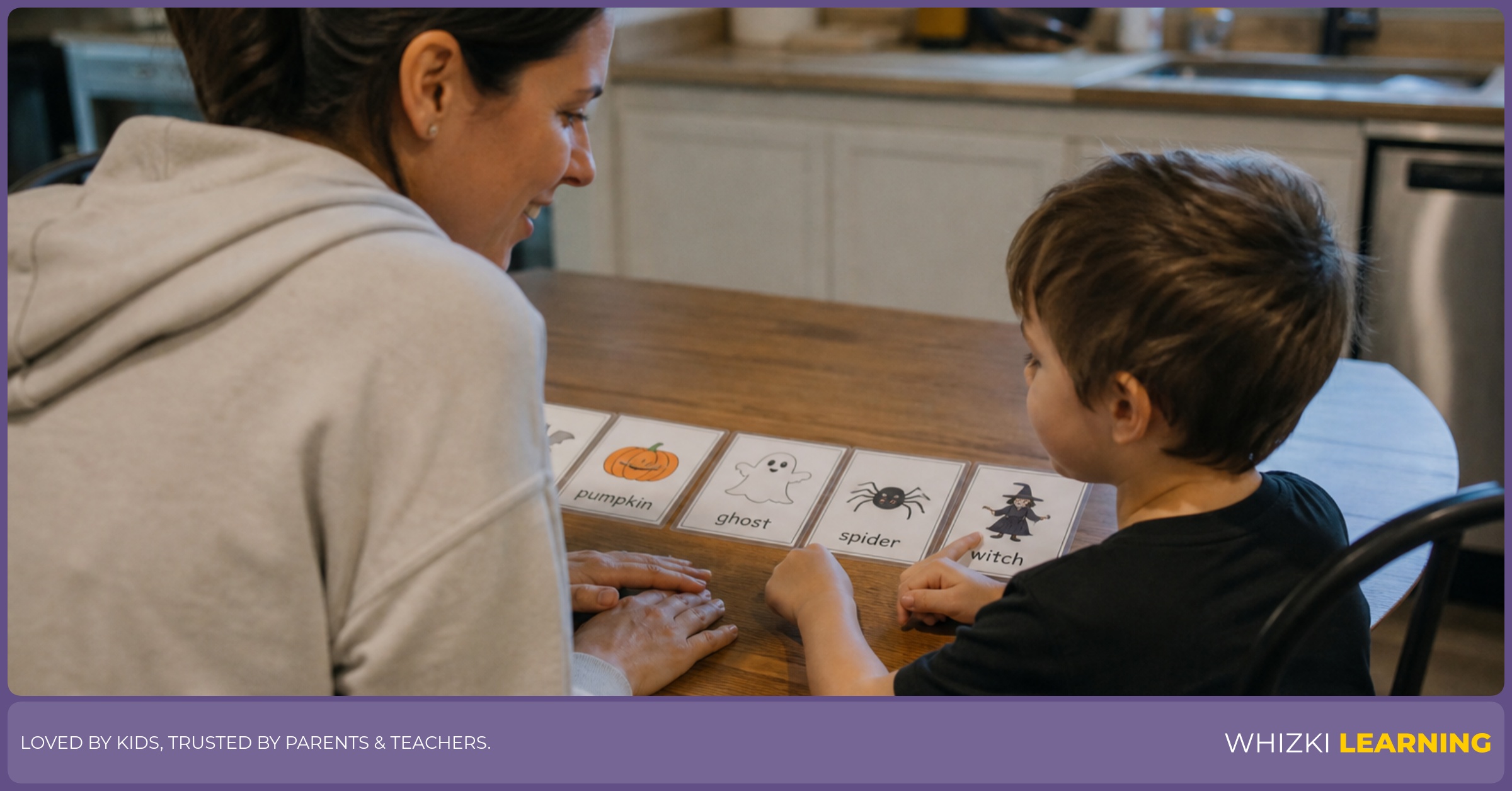 A parent helps a kindergarten child name friendly Halloween picture cards at a kitchen table.