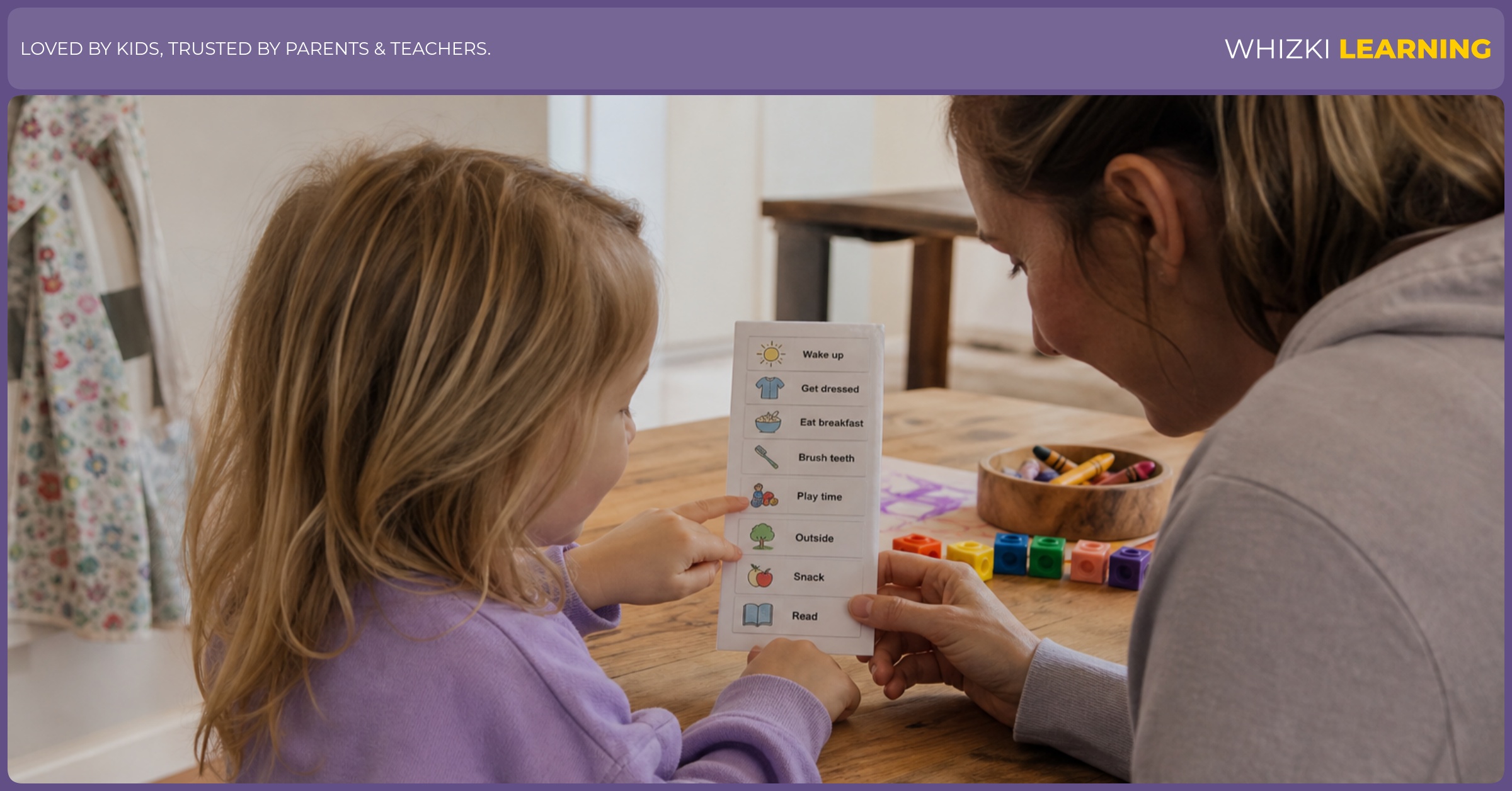 A parent sits beside a preschool child at a kitchen table while the child points to a small morning plan card with crayons nearby.