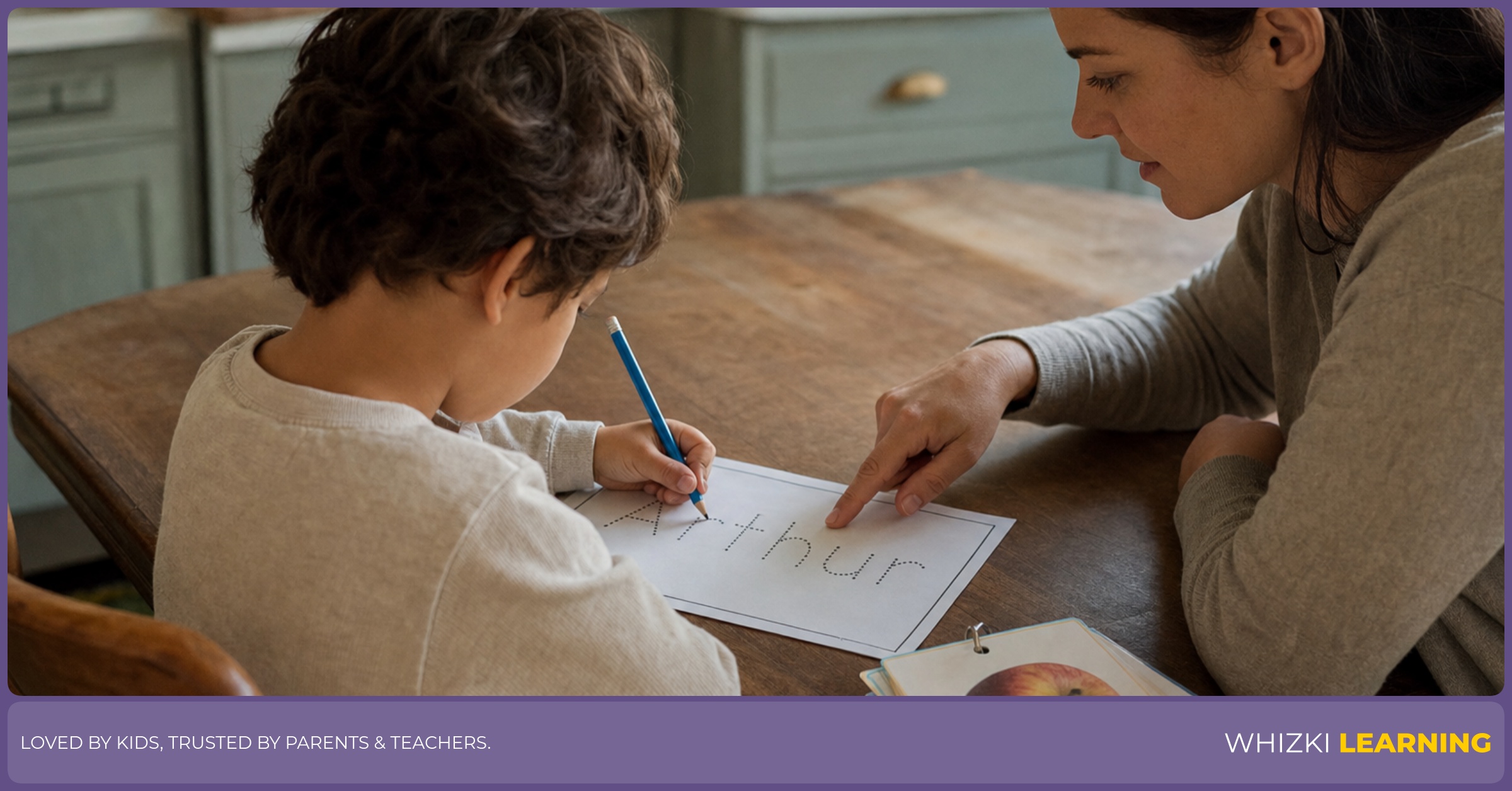 A parent sits beside a young child as the child traces a name worksheet with a pencil at the kitchen table.