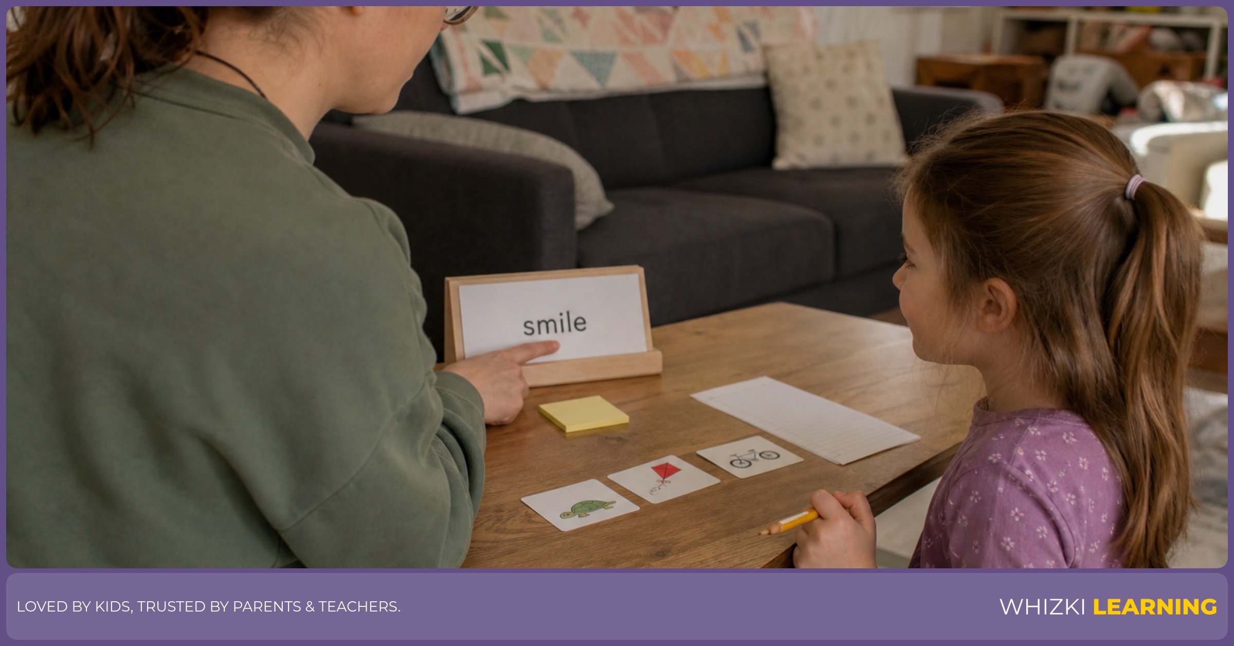 A caregiver and child practice reading a paper word card together with a pencil and sticky note on the table.