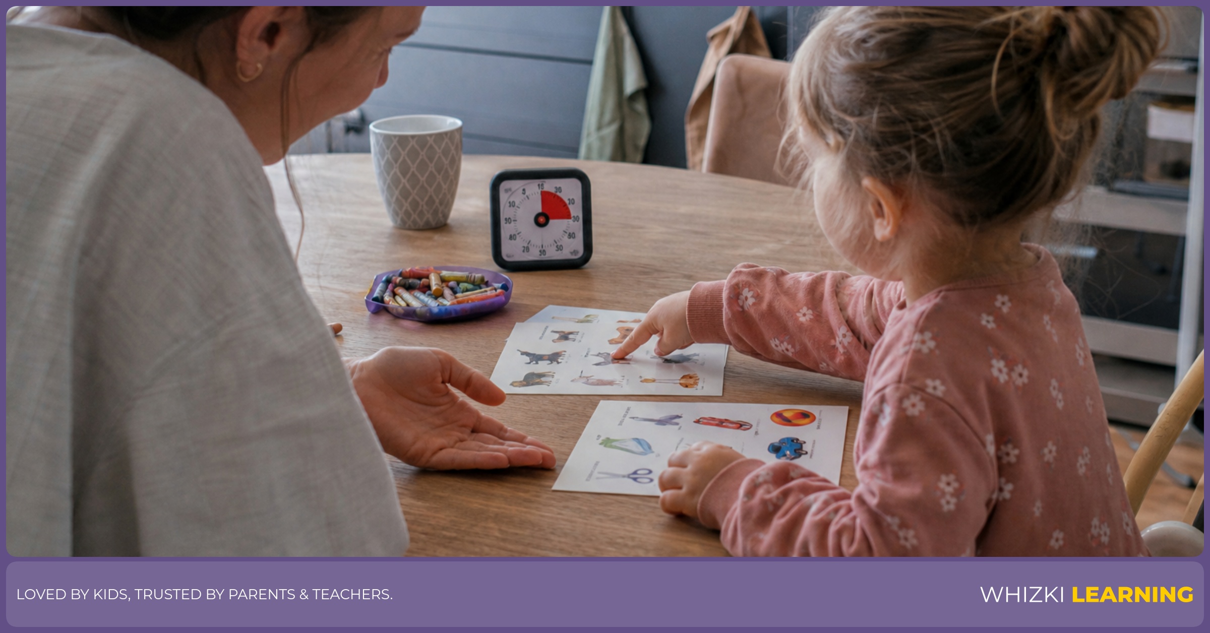 A caregiver sits beside a young child at a kitchen table while the child chooses between two paper worksheets.
