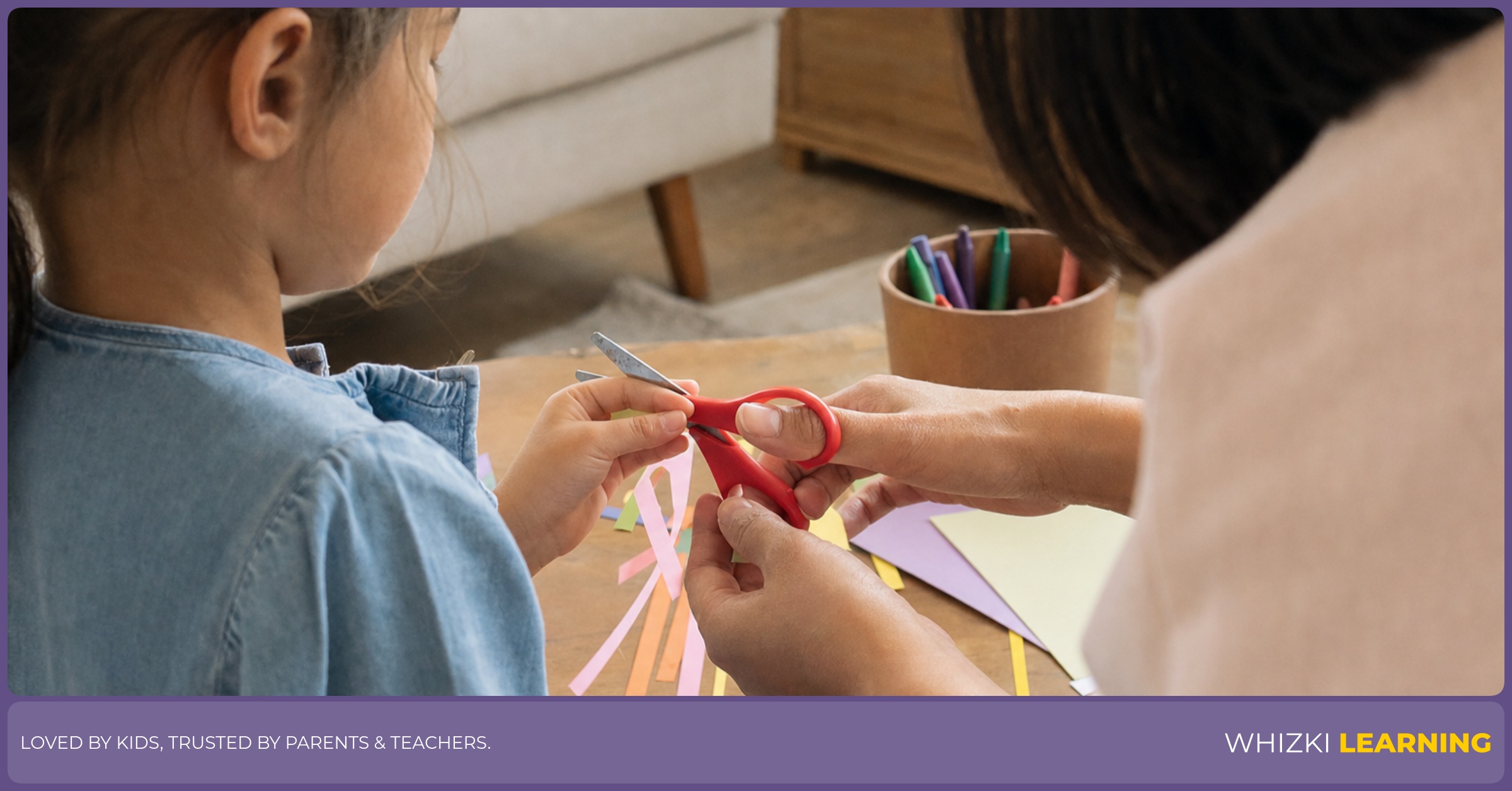 A parent gently guides a child’s scissor grip while paper strips and crayons sit on the table.