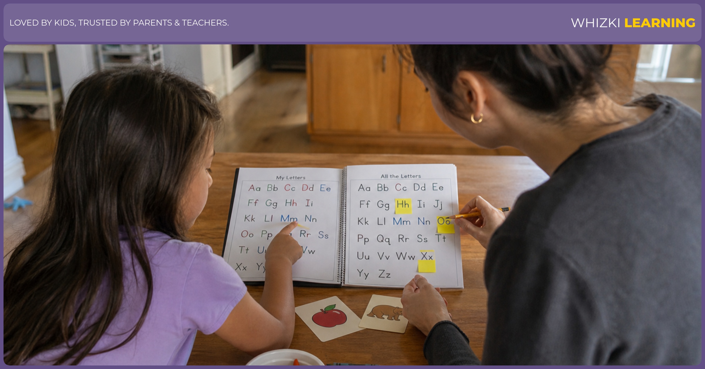 A parent sits beside a child comparing English and Ukrainian alphabet pages with sticky notes at a kitchen table.