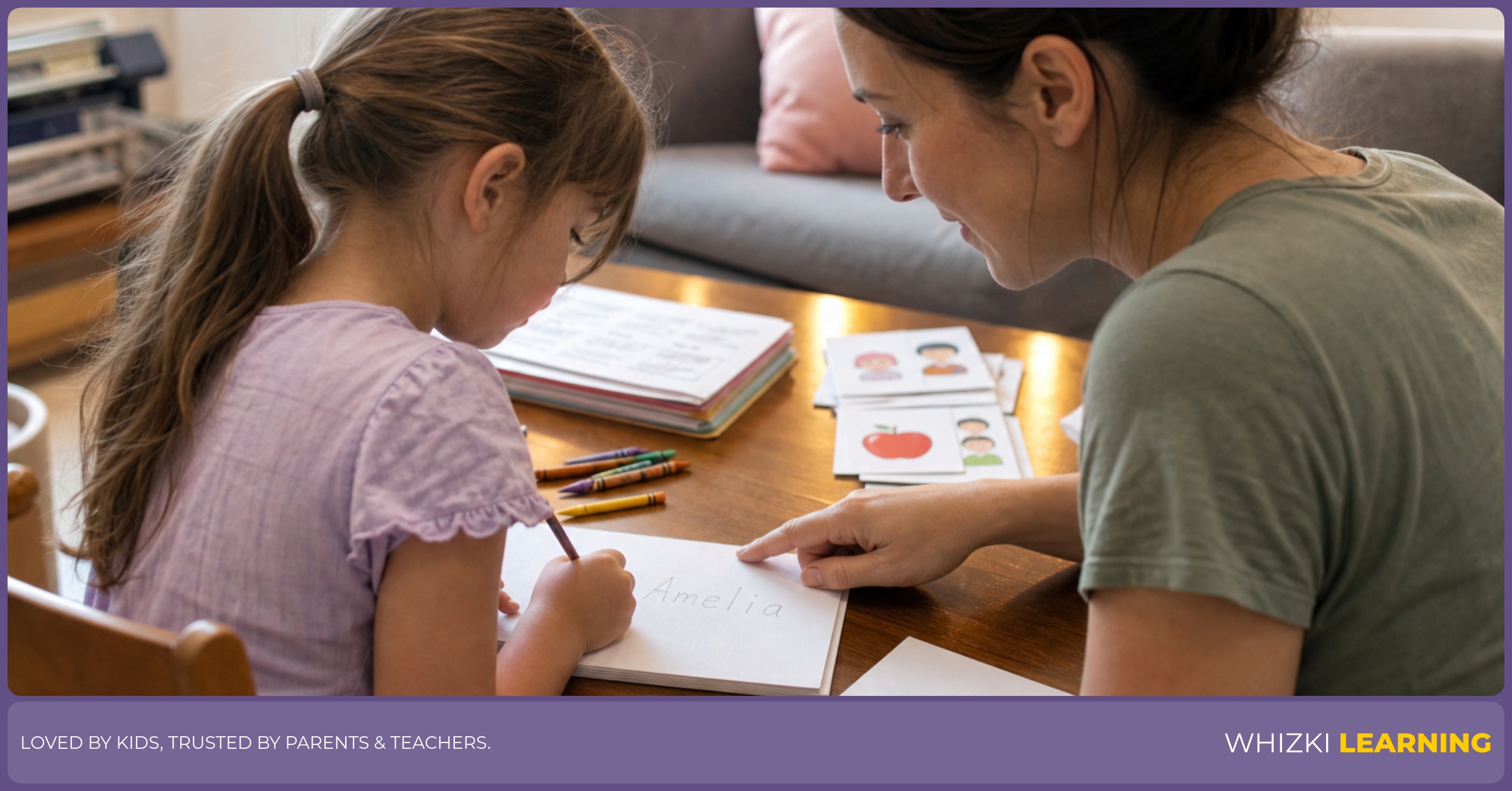 A caregiver sits beside a young child at a table while the child copies a first name onto paper with a short pencil.