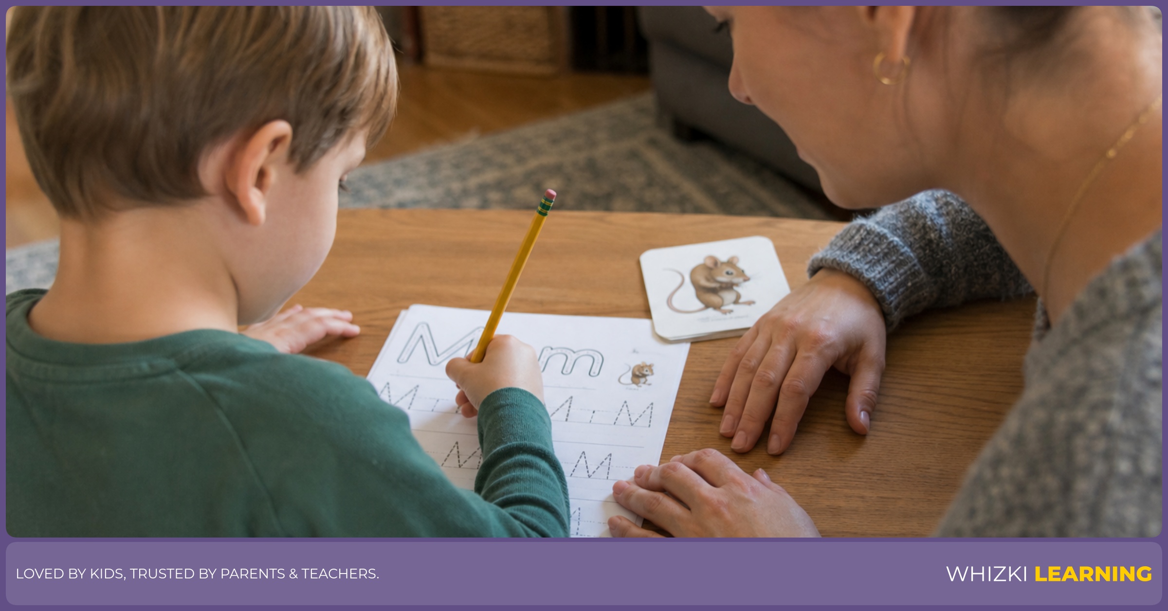A child traces an M verb on paper while an adult guides the pencil grip at the table.