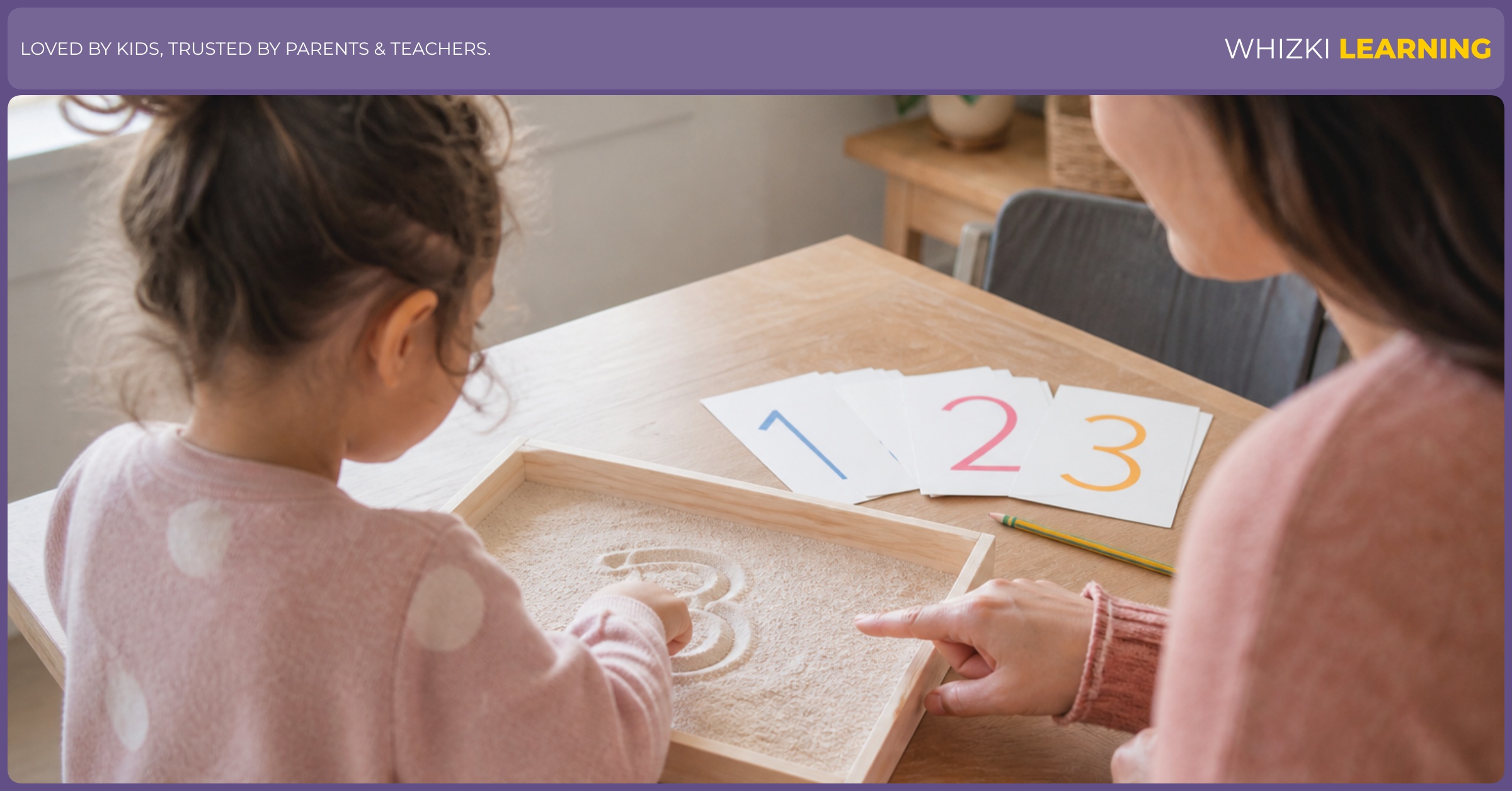A parent helps a young child trace a number in a shallow sand tray at the kitchen table.