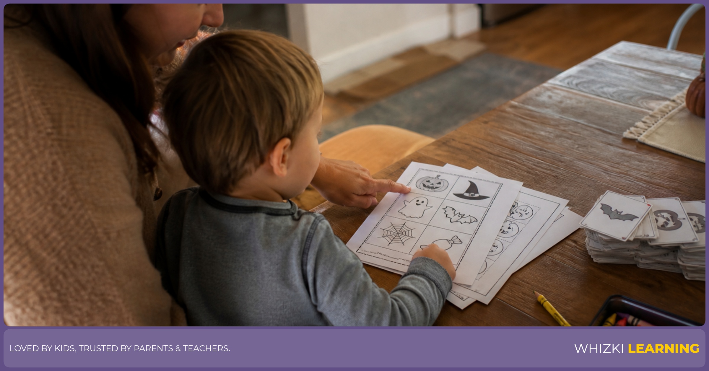 A parent and child sit at a kitchen table choosing one Halloween worksheet page with crayons and pencils nearby.