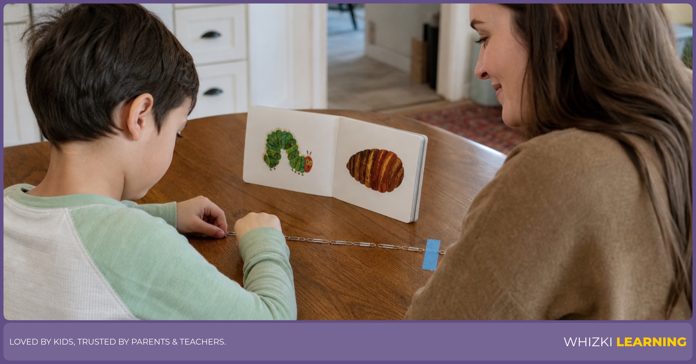 A parent helps a young child line up paper clips along the edge of a small book on a kitchen table.