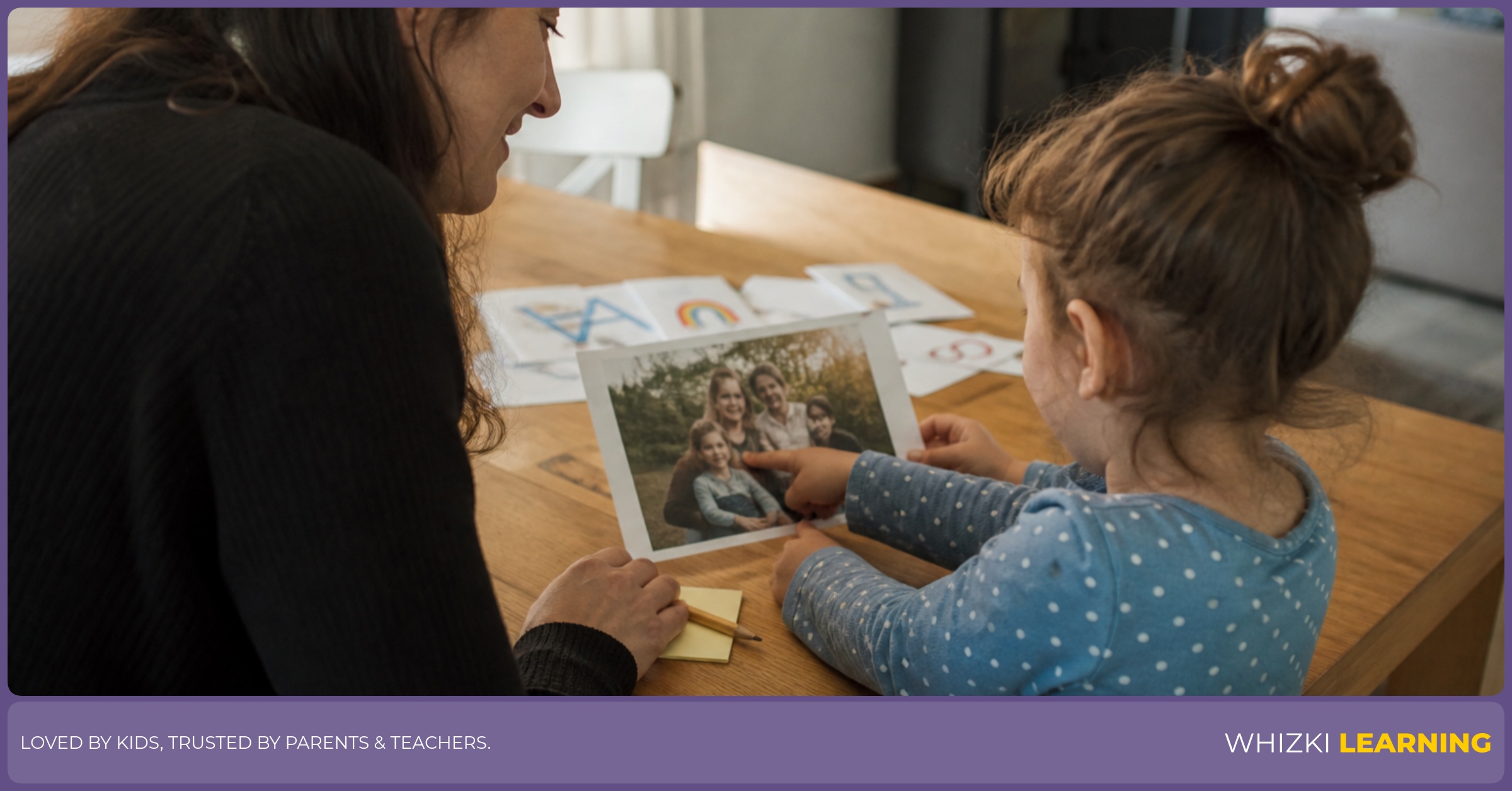 A parent and child sit at a kitchen table pointing to a printed family photo and talking together.