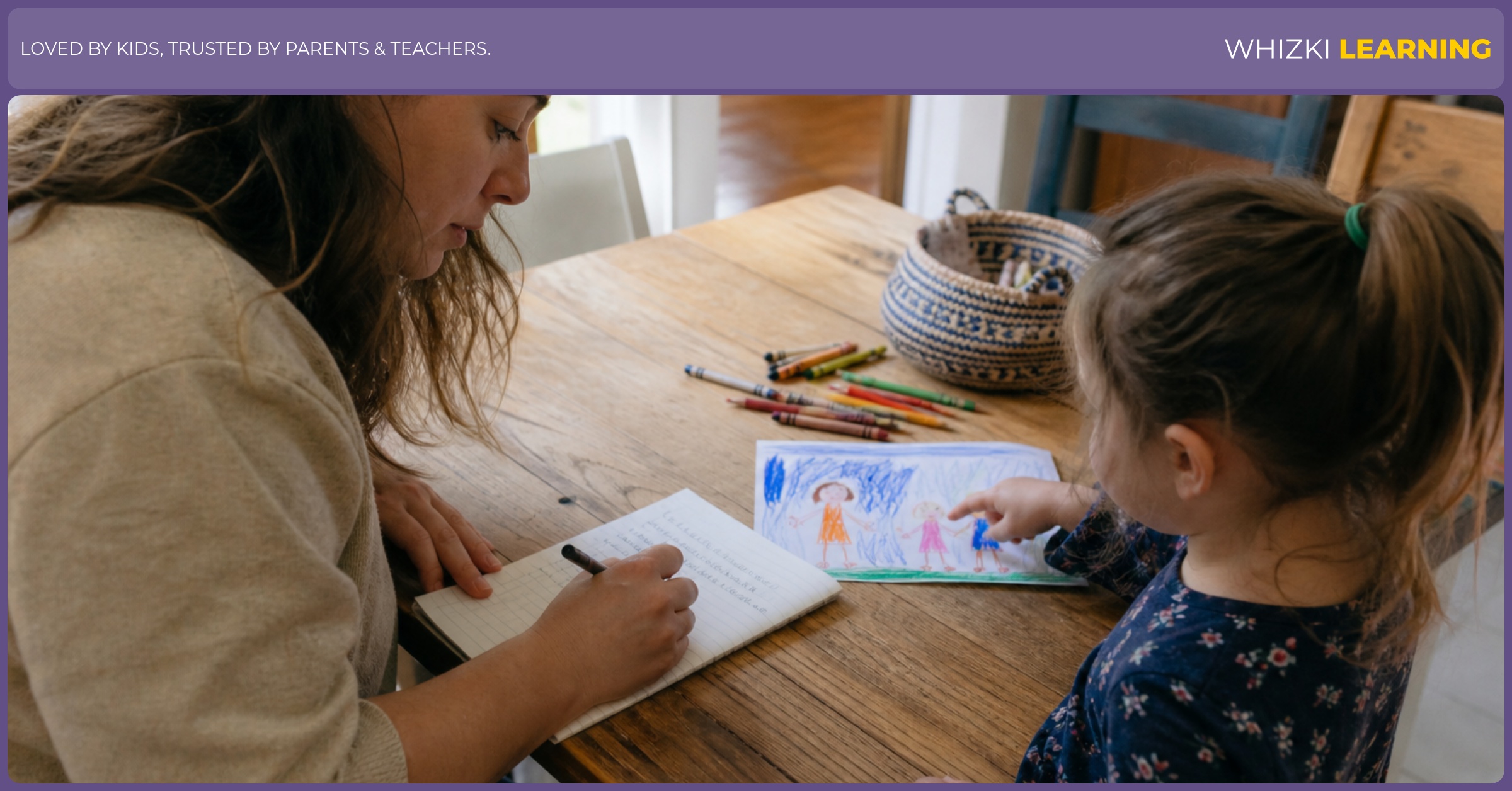 A parent writes a child’s sentence under a crayon drawing while the child points to the picture at a kitchen table.